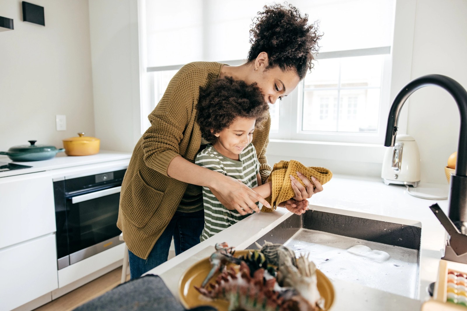 A parent and child washing dishes together at a stainless steel sink in a bright white kitchen